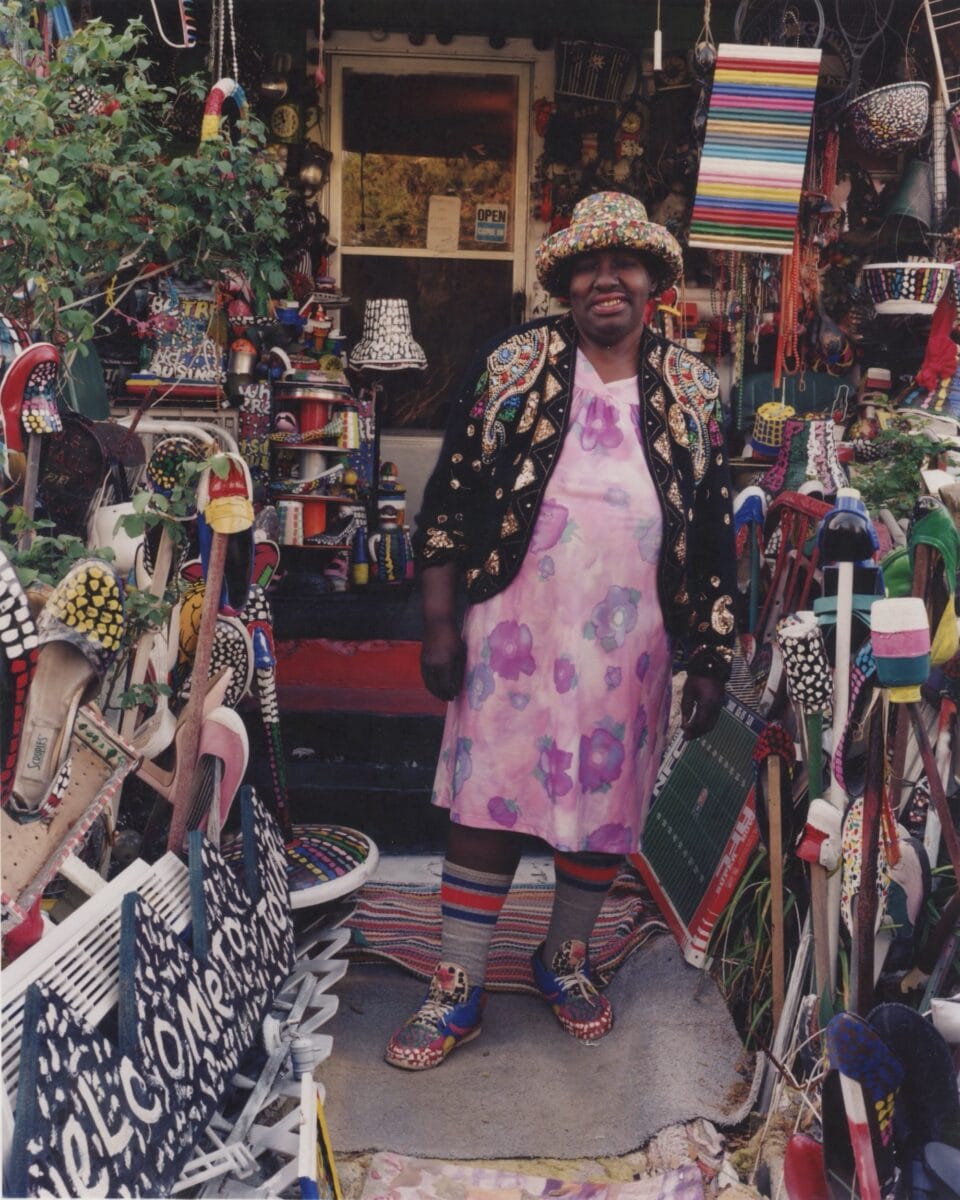 a Black woman in a pink and purple floral dress and hat poses for the camera in front of her home where the walkway is lined with found object assemblages