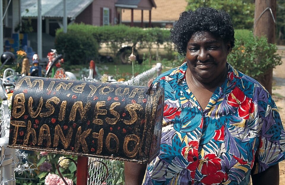 a Black woman in a floral shirt stands next to a mailbox painted with "mind your business. thank you"