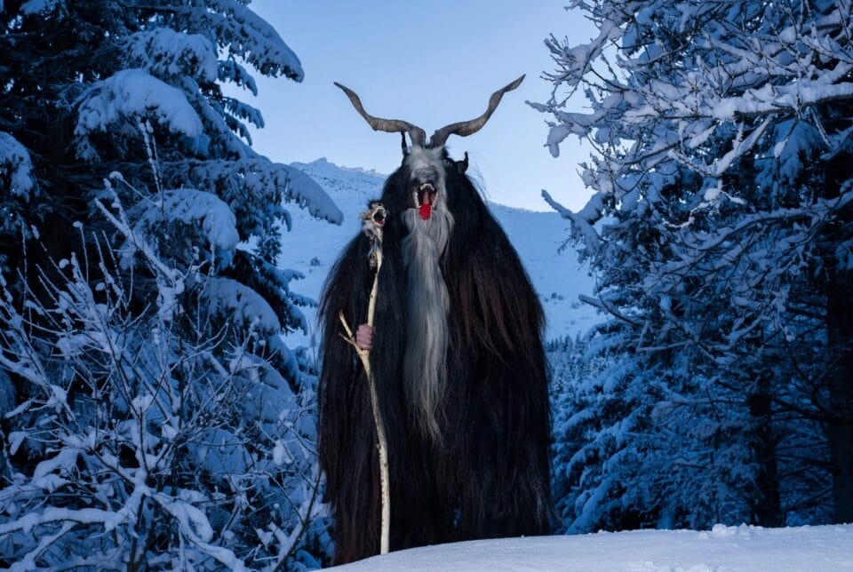 a figure wearing a scary costume made from goat hair and horns, holding a staff that has an open mouth on it that mimics the open, animalistic mouth of the mask