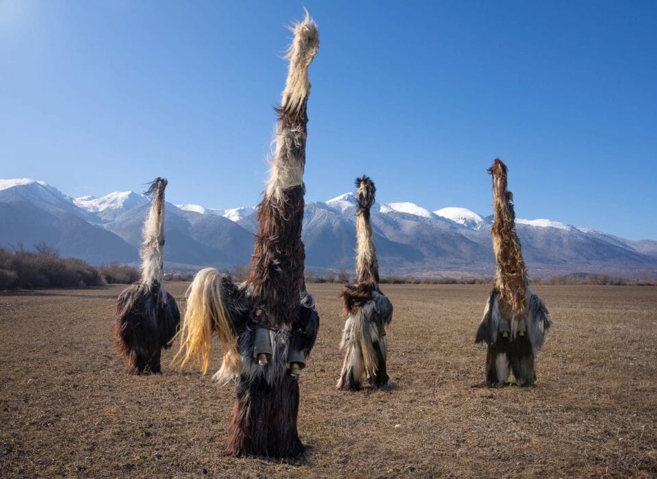 four costumed figures stand in a field in front of a mountain range in Bulgaria, all wearing furry outfits with very tall hats that make them appear like supernatural creatures