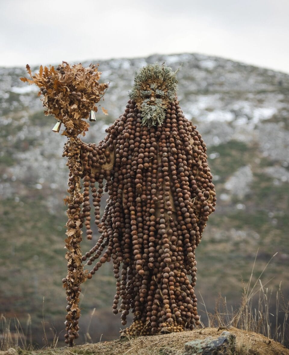 a figure stands on a hilltop in Spain, holding a staff covered in leaves and bells, and wearing an outfit completed covered in strands of nuts