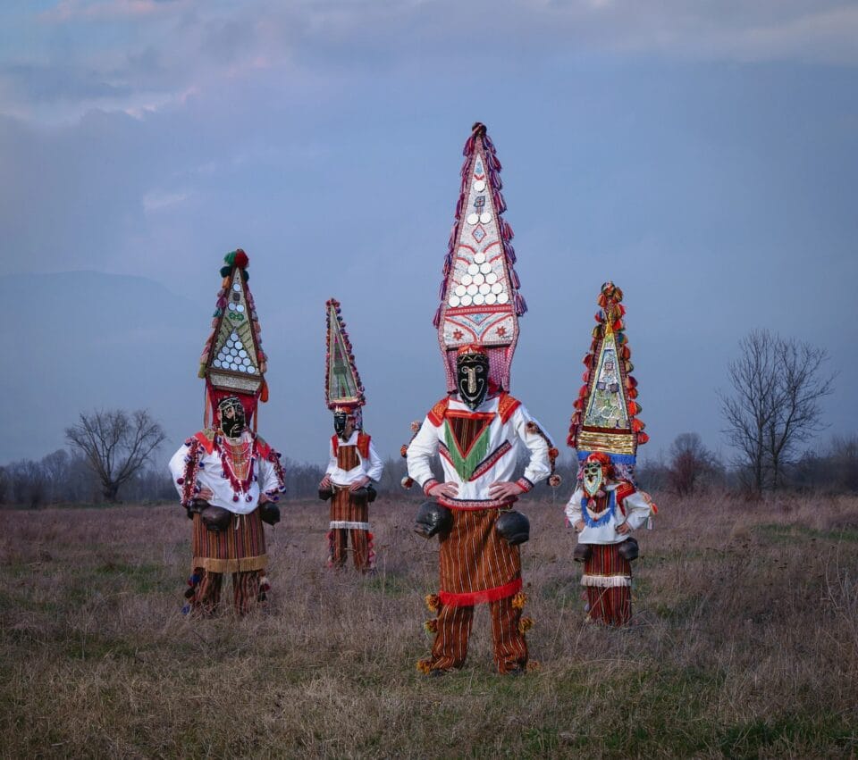 a group of four costumed figures standing in a field with elaborate triangular headdresses, masks, and colorful outfits